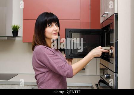 Una giovane donna guarda la macchina fotografica e toglie un piatto dal forno a microonde Foto Stock