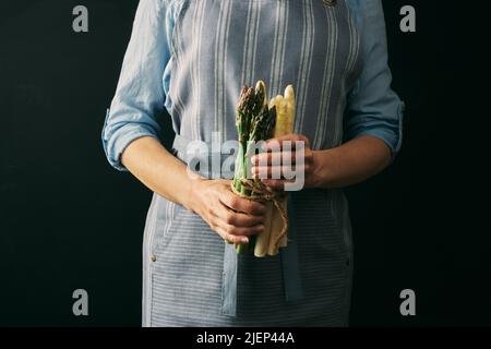 Coltivare la cuoca femminile senza volto in grembiule tenendo mix di asparagi crudi e sani su sfondo nero durante la preparazione della cottura in studio Foto Stock