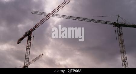 Sagome di gru edili e edificio residenziale incompiuto contro l'alba sopra la città. Edilizia abitativa, condominio costruzione di nuovi edifici residenziali. Sullo sfondo del cielo del tramonto. Foto Stock