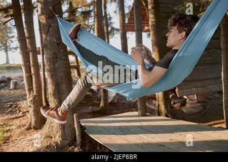 Bell'uomo che ha relax giorno in casa foresta. Sorseggiando caffè in amaca in legno al mattino. Foto Stock