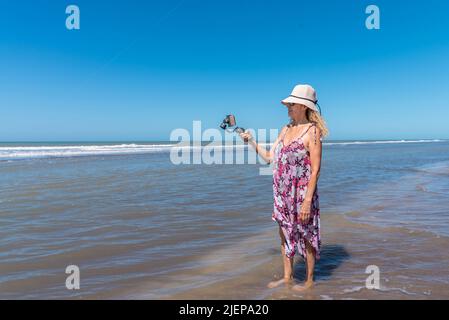 Donna bionda in estate vestire in piedi sull'acqua del mare usando un mobile attaccato su un gimbal. Foto Stock