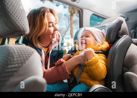 Ritratto di giovane madre sorridente allaccia la cintura di sicurezza sul seggiolino in cui è seduto il bambino piccolo felice. Il concetto di viaggio sicuro auto e guarire Foto Stock