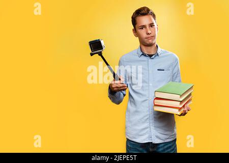 Il concetto di vlogging e di istruzione. Un ragazzo insoddisfatto in una maglietta blu tiene libri di testo e prende un selfie o un video su una fotocamera con un monopo Foto Stock