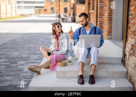 I colleghi di stile lavorano su un computer portatile e mangiano cibo da portare via in una strada all'aperto Foto Stock