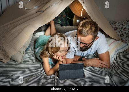 Ritratto di un ragazzo di 6 anni e suo padre che si divertono a giocare in tenda teepee. Padre e figlio usano un tablet digitale per guardare cartoni animati o giocare a giochi per computer che si trovano nella tenda del bambino a casa. Foto Stock