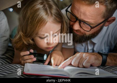 Ritratto di un ragazzo di 6 anni e suo padre che legge un libro in tenda teepee. Padre e figlio con libro di lettura torcia sotto coperta a casa. Foto Stock