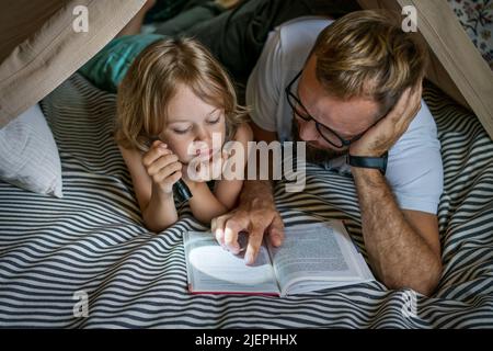 Ritratto di un ragazzo di 6 anni e suo padre che legge un libro in tenda teepee. Padre e figlio con libro di lettura torcia sotto coperta a casa. Foto Stock