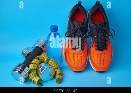 Sneakers arancioni, manubri, nastro di misurazione e bottiglia di acqua potabile in plastica su sfondo blu Foto Stock