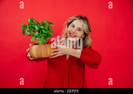 Donna bionda sorridente che sorride la pianta di basilico su sfondo rosso in studio, sorridendo il toothy sorriso, guardando la macchina fotografica Foto Stock