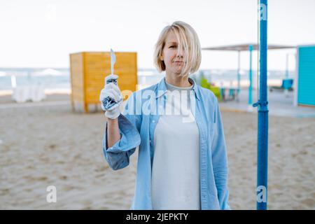 Ritratto di donna di mezza età con capelli discrete indossando T-shirt grigia, camicia blu, guanti da lavoro in cotone, in piedi sulla spiaggia sabbiosa, alzando la mano con pl bianco Foto Stock