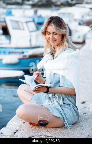 Felice giovane donna in denim azzurro jumpsuit è seduta sul molo con libro in mano, accanto a un bicchiere di vino rosso. Foto Stock