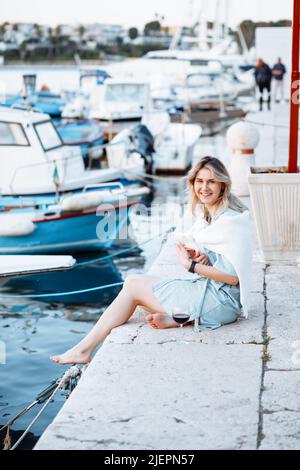 Vacanza al mare. Sorridente bionda si rilassa seduto sul molo con il libro in mano, c'è un bicchiere di vino accanto a lei. Foto Stock