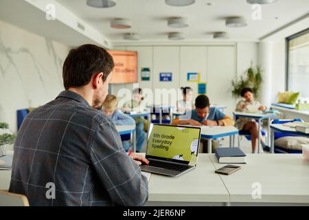 Vista posteriore a insegnante di sesso maschile utilizzando un computer portatile alla scrivania in aula con gruppo di bambini che studiano in background, spazio copia Foto Stock