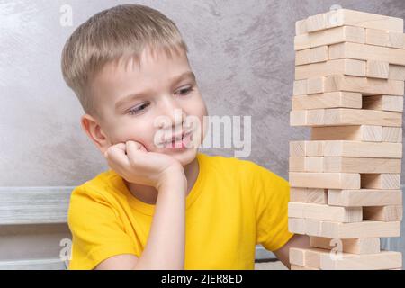 Interessato ragazzo biondo felice giocare gioco di bordo prendendo mattoni da torre di legno mantenere equilibrio avendo divertimento insieme a casa. Foto Stock