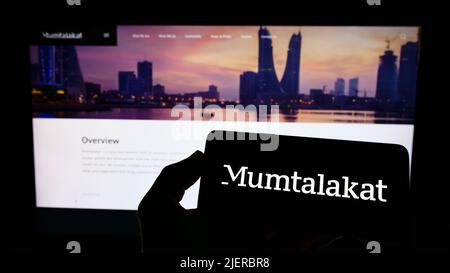 Persona che tiene il telefono cellulare con il logo della Bahrain Mumtalakat Holding Company sullo schermo di fronte alla pagina web aziendale. Mettere a fuoco sul display del telefono. Foto Stock