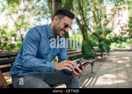 Messaggi di testo per uomo d'affari sorridenti sullo smartphone. Giovane imprenditore maschio in rete mentre si siede su panchina. Indossa i formals al parco in città. Foto Stock