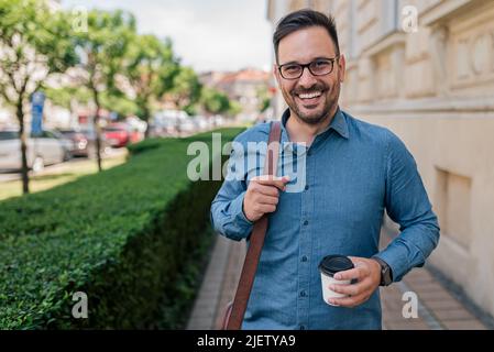 Ritratto di sorridente professionista con tazza di caffè sul marciapiede da piante. Il penduter maschio felice sta trasportando la borsa del laptop. Indossa i formals in città. Foto Stock