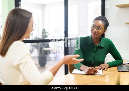 Donna d'affari fiduciosa, direttore esecutivo, ceo che parla con una collega afro-americana, che discute di strategia. Diverse donne partner brainstorming in sala riunioni, donna multirazziale prende appunti Foto Stock