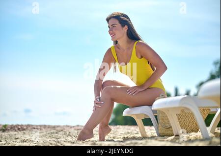 Signora allegra seduta sulla sdraio utilizzando la lozione solare Foto Stock