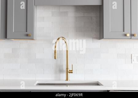 Un dettaglio lavandino della cucina girato con un rubinetto d'oro, backsplash in marmo, armadi grigi, e metallo dorato. Foto Stock