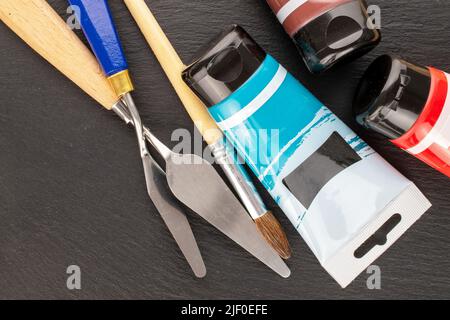Due coltelli per palette in metallo, una spazzola e vernici su una pietra ardesia, primo piano, vista dall'alto. Foto Stock