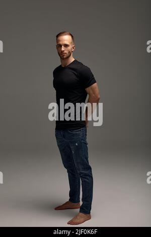 Un attraente uomo bianco in jeans e camicie nere si abbonde in piena crescita su sfondo grigio. Spazio di copia Foto Stock