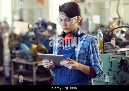 Donna brutale giovane seria in occhiali protettivi e tuta blu in officina e lettura delle specifiche tecniche sul tablet Foto Stock