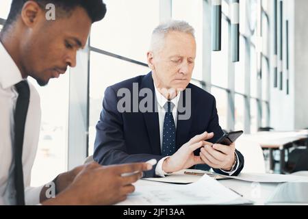 Uomo d'affari senior concentrato in giacca formale seduta al tavolo con il collega e controllare il messaggio al telefono durante la riunione Foto Stock
