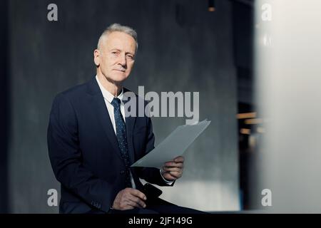 Ritratto di dirigente senior serio e rigoroso con capelli grigi seduti nella lobby scura e in possesso di documenti aziendali Foto Stock