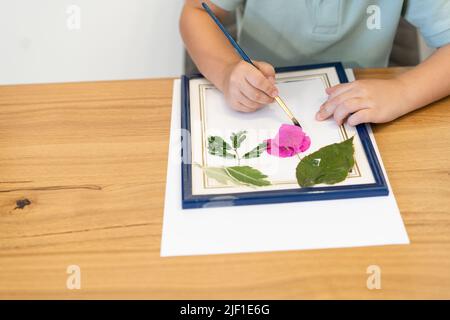 Mani di bambino con album, foglie e fiori che fanno herbarium su tavola di legno da pranzo in casa. Foto Stock