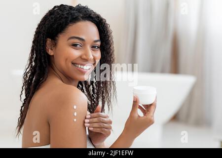 Concetto di self-Care. Giovane donna Nera che applica lozione idratante corpo sulla pelle Foto Stock