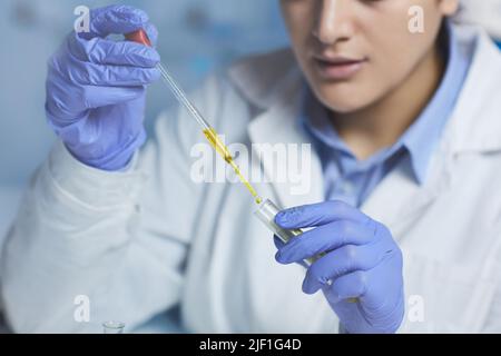 Primo piano di una scienziata femminile concentrata in guanti sterili e camice da laboratorio con contagocce durante la miscelazione dei reagenti nella provetta del campione medico Foto Stock