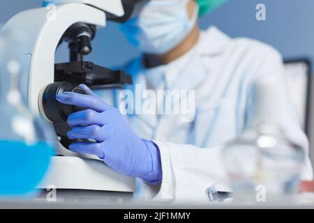 Primo piano di un biologo irriconoscibile in guanti di gomma e cappotto bianco seduto alla scrivania e microscopio di regolazione in laboratorio Foto Stock