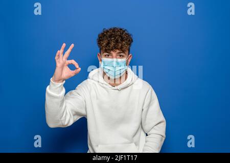 Bell'uomo caucasico che mostra il gesto giusto. La foto di un uomo sano indossa una maschera protettiva contro le malattie infettive e l'influenza. Concetto di assistenza sanitaria Foto Stock