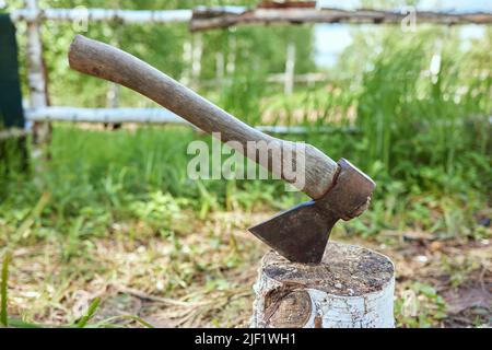 Un'ascia vecchia grande sporge da un blocco di trinciatura del ceppo di legno sullo sfondo di una radura verde di trifoglio in una giornata di sole di estate Foto Stock