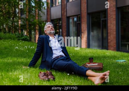 Un uomo d'affari maturo che riposa e siede a piedi nudi nel parco, sentendosi libero, fuggendo dal lavoro, concetto di equilibrio di vita di lavoro. Foto Stock