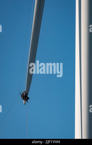 Due uomini in imbracature di sicurezza lavorano su una lama di mulino a vento sfumata mentre sospeso dall'alto. Foto Stock