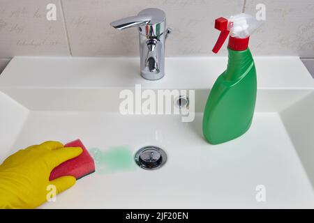 Mano in guanti protettivi gialli pulisce il lavandino in bagno con una spugna e detergente Foto Stock