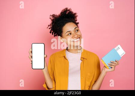 Felice eccitata giovane afroamericana, pronta per le vacanze, tiene il passaporto con i biglietti in mano, e telefono con schermo bianco vuoto, stand su isolato sfondo rosa, guardando gioiosamente da parte, sorriso Foto Stock