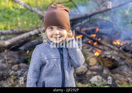 Primo piano ritratto di un ragazzo sorridente di quattro anni che indossa berretto da beanie e giacca blu con zip in piedi nella natura. Fuoco di camping sfocato sullo sfondo. Foto Stock