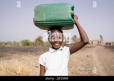 Sorridente ragazza di villaggio africano che porta un grande barattolo verde sulla sua testa come lei cammina alla stazione di acqua della comunità; diritto umano di bere sicuro wat Foto Stock