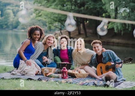 Gruppo di amici che si divertono al picnic vicino a un lago, seduti su coperta mangiare e suonare la chitarra. Foto Stock