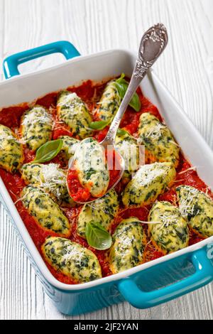 Malfatti, gnocchi di ricotta di spinaci italiani in salsa di pomodoro con erbe e parmigiano grattugiato in un piatto di cottura in ceramica con cucchiaio Foto Stock