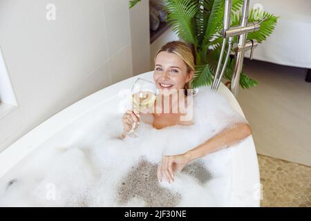 Giovane donna piuttosto bionda sorridente, sdraiata in bagno con bollicine e vino bianco. Bagno. Foto Stock