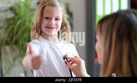 Il medico ascolta il bambino con stetoscopio, la ragazza tiene i pollici in su Foto Stock