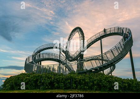 Scultura di montagne russe pedonali Tiger & Turtle su Magic Mountain con alcune persone in movimento sfocate contro un cielo nuvoloso tramonto, installazione d'arte e l Foto Stock