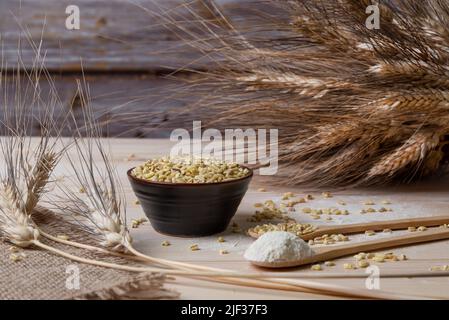 Ciotola piena di semi di grano tenero e di spighe secche di grano insieme ad un cucchiaio da tavola di farina di grano. Stadi diversi di grano Foto Stock