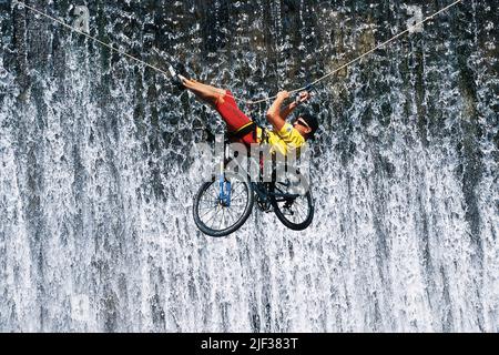 Mountainbiker con la sua bicicletta attraversando un canyon appeso a una fune di arrampicata, cascata in background Foto Stock