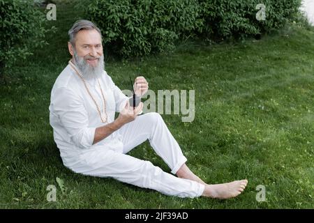 allenatore di yoga senior felice seduto con diffusore e bastoncini di aroma sul prato verde Foto Stock