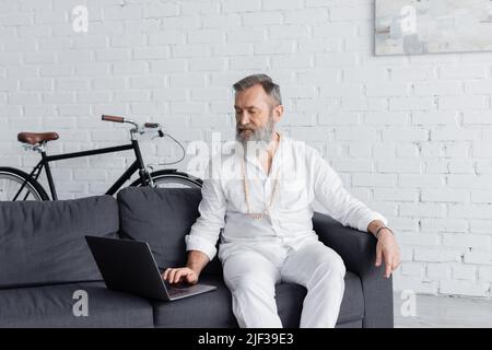 allenatore senior per la mente dei maestri in abiti bianchi utilizzando un computer portatile mentre si siede sul divano a casa Foto Stock